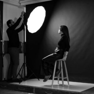 A photographer adjusts studio lighting for a female model seated on a stool.