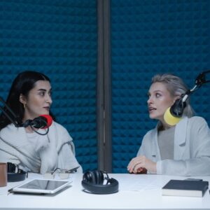 Two women engaged in conversation, recording podcast in studio with microphones and headphones.